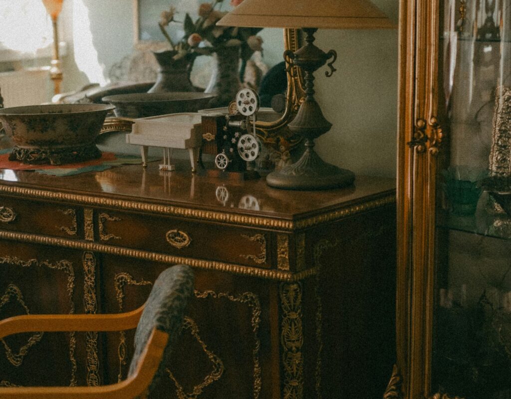 Antikt skrivbord eller byrå i mörkt trä med förgyllda beslag och dekorativa detaljer. På möbeln står flera äldre föremål, bland annat en liten dekorativ filmprojektor, en miniatyrflygel, metallskålar samt en klassisk bordslampa med tygskärm. Intill syns en stol med träarmstöd och i bakgrunden anas en glasmonter med fler antikviteter, vilket skapar en tidstypisk och samlad miljö.