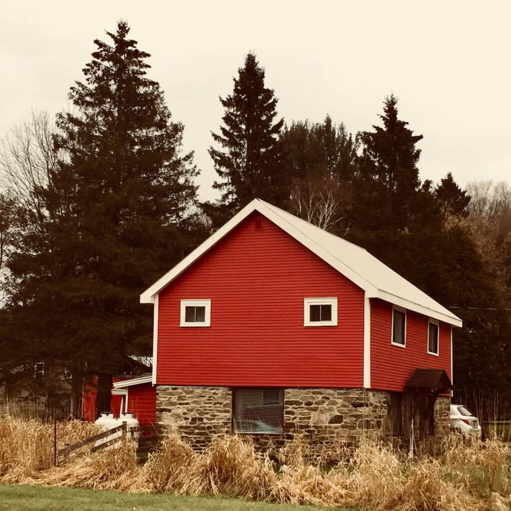Röd träbyggnad med vitmålade knutar och sadeltak, uppförd på en grund av natursten. Huset är omgivet av högt gräs och granar i en lantlig miljö, vilket ger ett traditionellt svenskt uttryck med koppling till äldre bebyggelse och landsbygd.