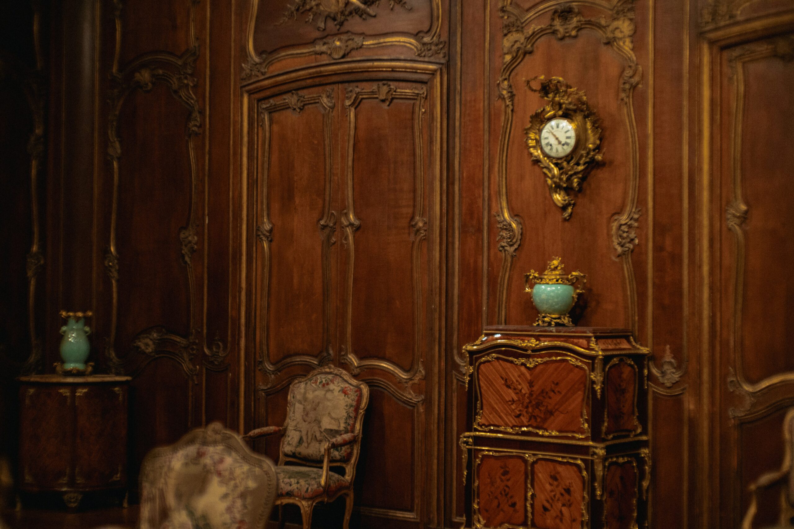 Interiör med rikt dekorerade träpaneler och antika möbler i rokokostil. I rummet syns en utsirad byrå med förgyllda detaljer, en väggmonterad pendylklocka, stoppade stolar samt dekorativa urnor, vilket skapar en elegant och historiskt präglad miljö.