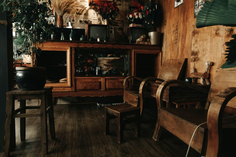 Interiör med äldre träinredning bestående av vitrinskåp, stolar och småbord i patinerat trä. Rummet är fyllt med föremål som keramik, växter och äldre elektronik, vilket skapar en varm och genuin miljö med tydlig känsla av samling, hantverk och äldre bruksmiljö.