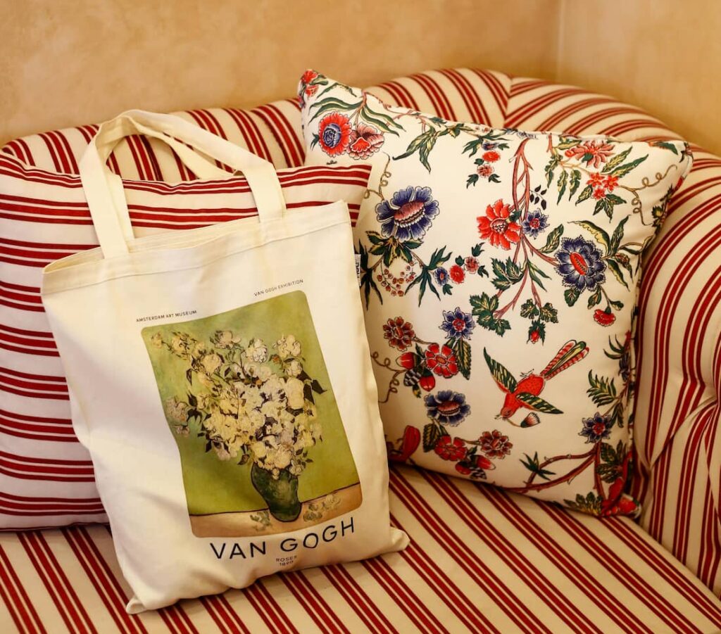 Randig stoppad soffa i rött och vitt med dekorativa kuddar, varav en med färgstarkt blommönster. På soffan ligger en tygkasse med tryck av ett blomsterstilleben av Van Gogh, vilket skapar en varm och personlig inredningsmiljö med konstnärlig prägel.