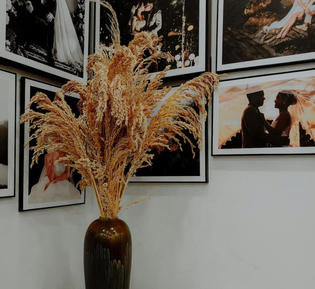 Stor golvvas i mörk keramik fylld med torkade prydnadsgräs, placerad framför en vägg med inramade fotografier i svartvita och varma toner. Kombinationen av naturmaterial och konst skapar en stilren och harmonisk inredningsmiljö med galleri­känsla.