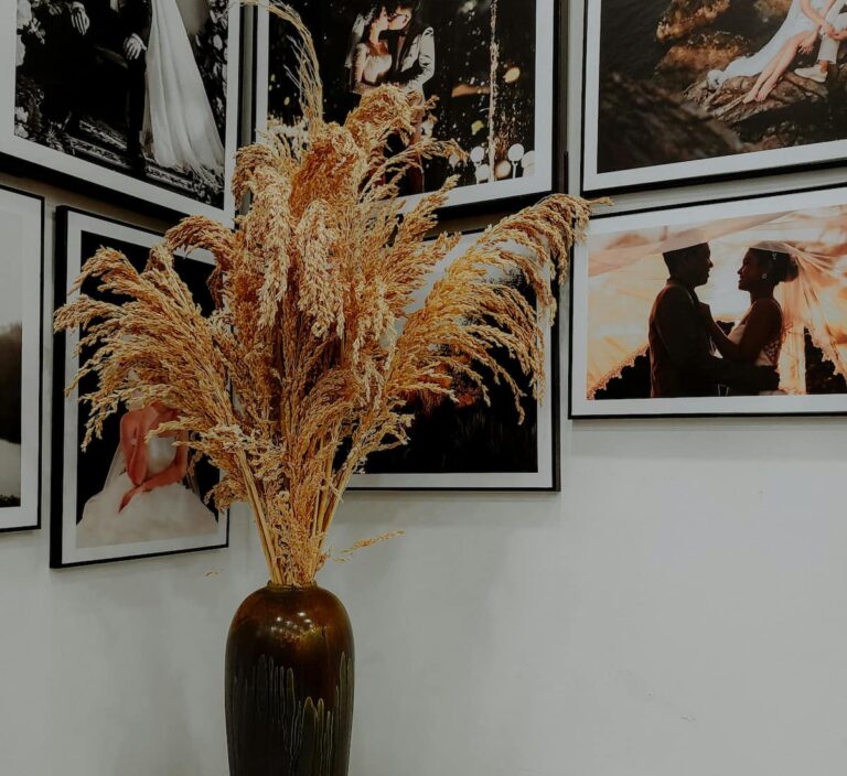 Stor golvvas i mörk keramik fylld med torkade prydnadsgräs, placerad framför en vägg med inramade fotografier i svartvita och varma toner. Kombinationen av naturmaterial och konst skapar en stilren och harmonisk inredningsmiljö med galleri­känsla.