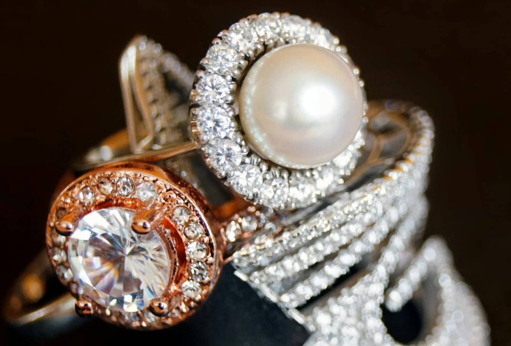 Närbild av flera ringar i guld och vitmetall, dekorerade med diamanter och en central pärla. Smyckena har detaljrik infattning och gnistrande stenar, vilket ger ett exklusivt och tidlöst uttryck som förknippas med värdefulla smycken.